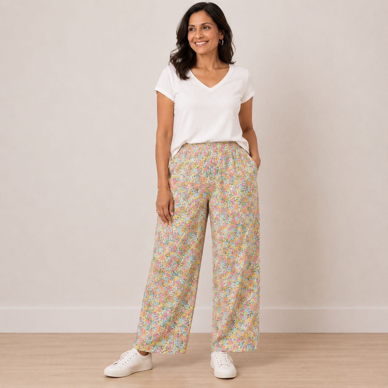 A woman modeling a pair of wide-leg pants with a dense pastel floral print, paired with a white V-neck t-shirt and white sneakers, standing against a neutral beige wall.