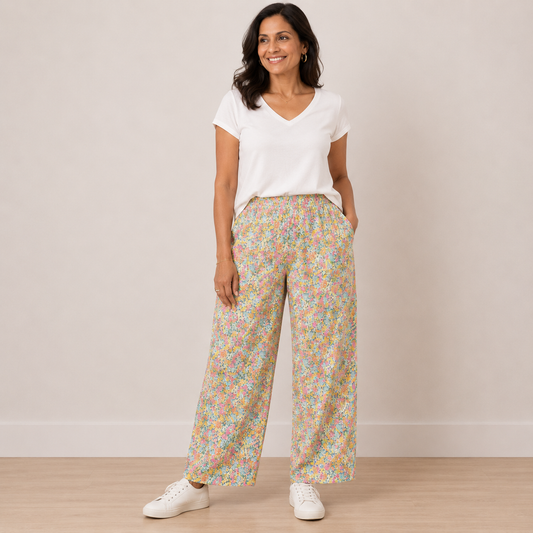 A woman modeling a pair of wide-leg pants with a dense pastel floral print, paired with a white V-neck t-shirt and white sneakers, standing against a neutral beige wall.