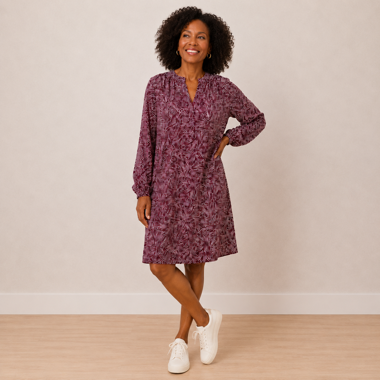 A woman modeling a long-sleeve, knee-length dress with a purple and pink abstract floral pattern, paired with white sneakers, standing against a neutral beige wall and light wood floor.