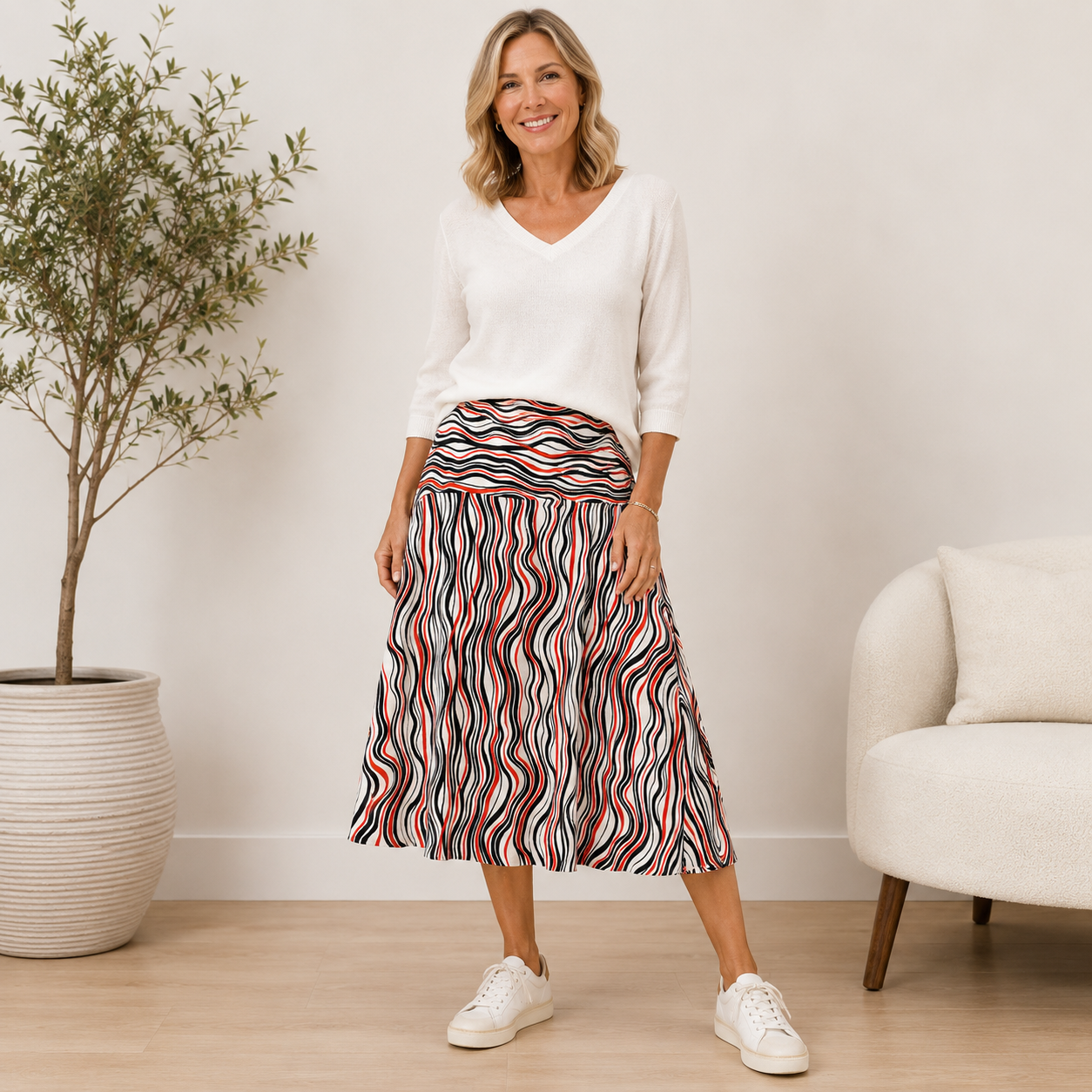 A woman stands in a bright indoor room wearing a white V-neck long-sleeve top and a high-waisted midi skirt with a wavy multicolor stripe pattern in red, blue, black, and white. She is wearing white sneakers and smiling.