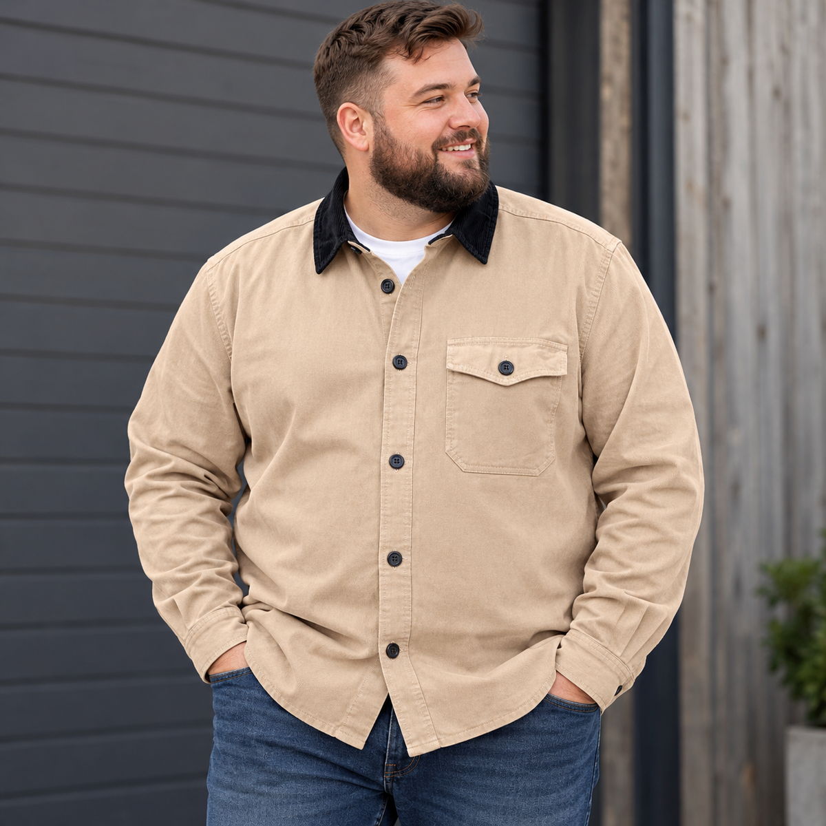 A man wearing a beige button-up overshirt with a black corduroy collar, styled with blue jeans over a white t-shirt, standing in front of a dark gray wooden wall.