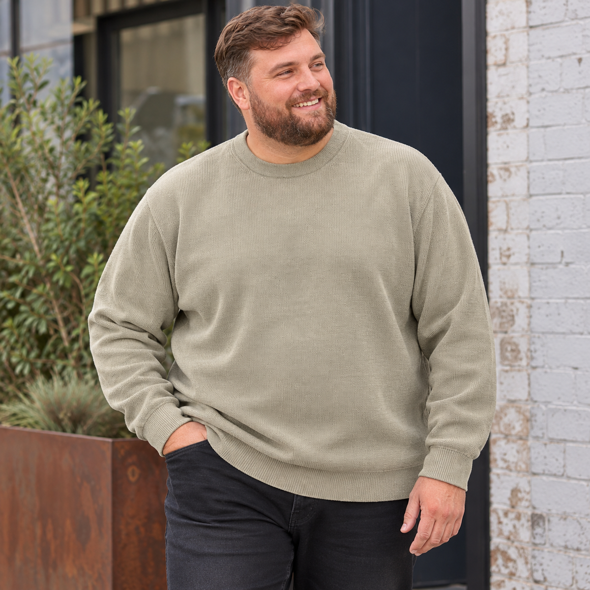 A smiling man wearing a light sage green crewneck sweatshirt and dark jeans, standing outdoors in front of a white brick wall and a planter box.