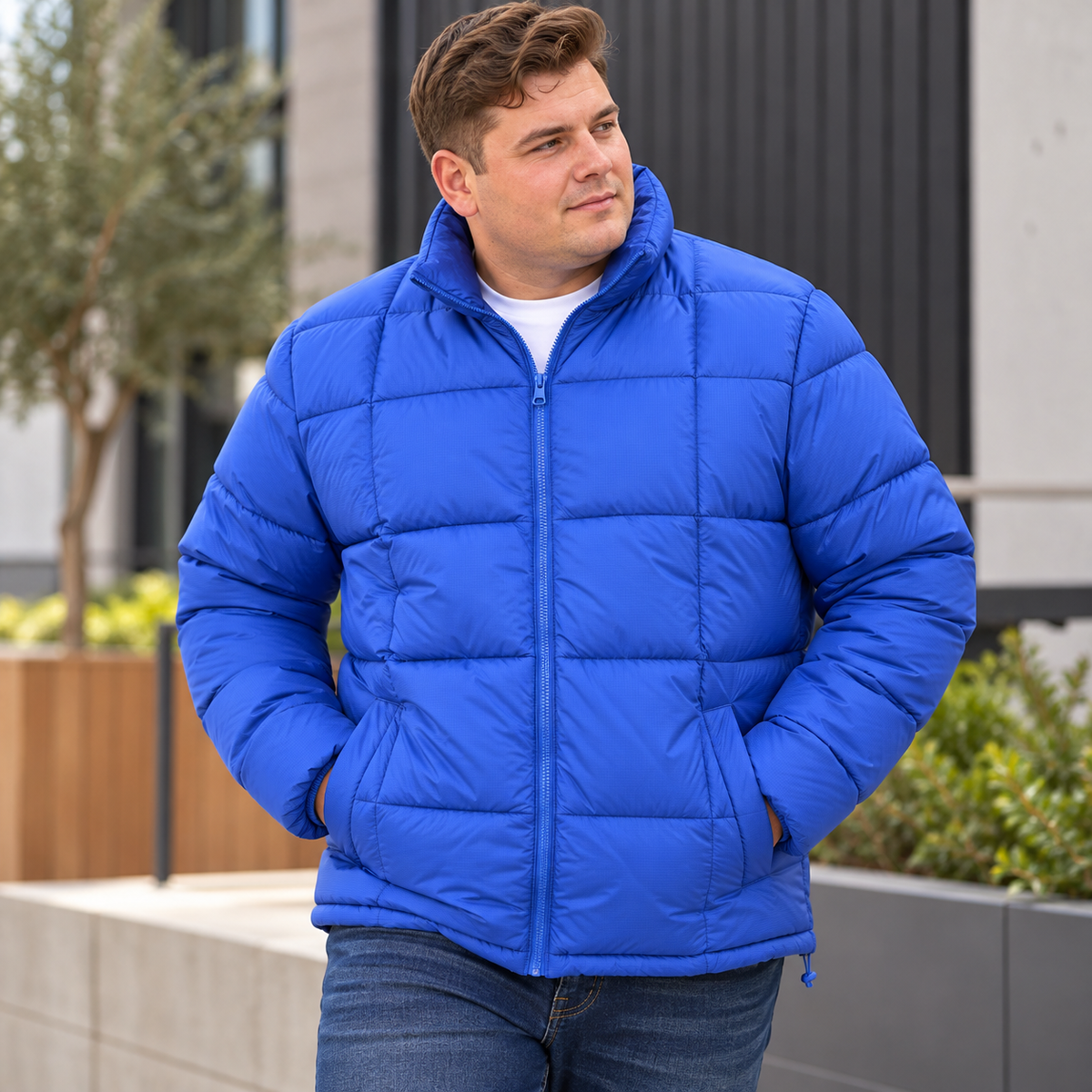 A man wearing a bright blue puffer jacket over a white t-shirt and blue jeans, standing outdoors in front of a modern building with planters.