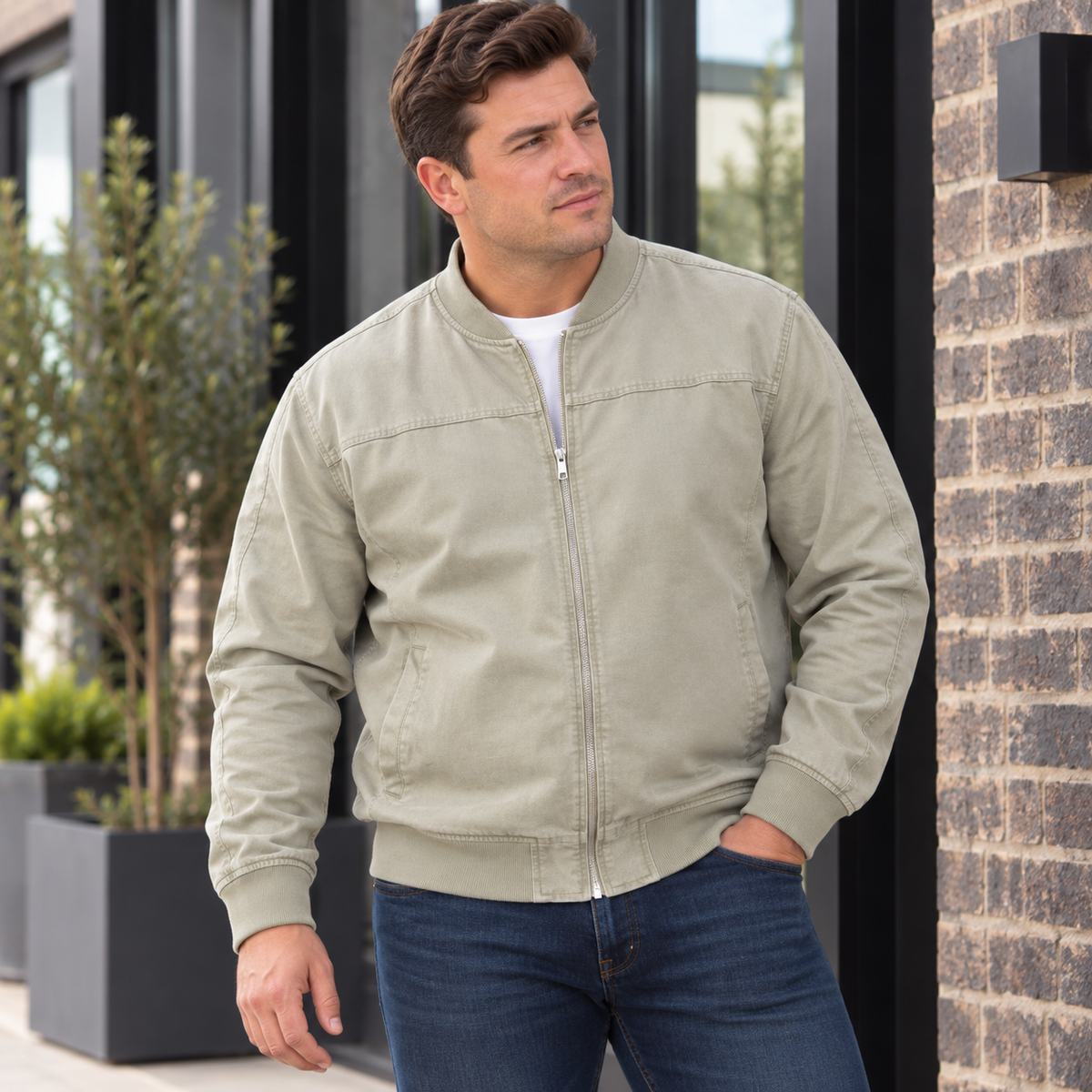 A man wearing a light khaki zip-up bomber jacket over a white t-shirt and blue jeans, standing outdoors next to a brick wall.