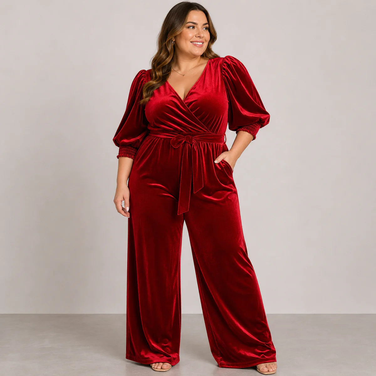 A woman modeling a deep red velvet jumpsuit with puff sleeves, a V-neckline, and a self-tie waist belt, set against a neutral studio background.