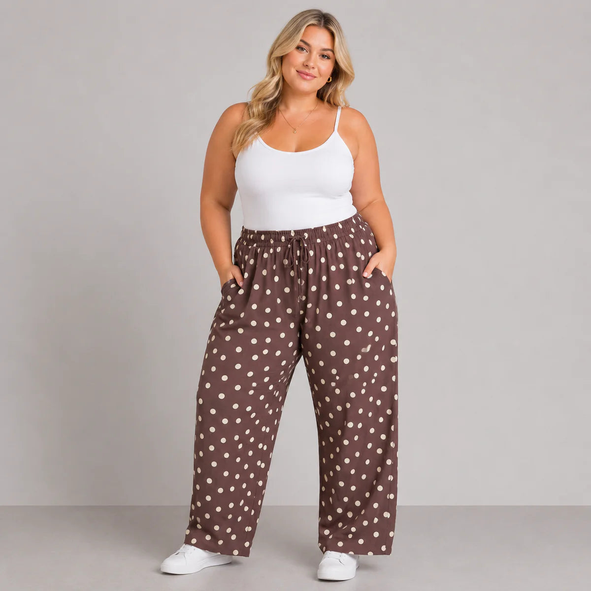 A woman modeling wide-leg brown pants with a white polka dot pattern, paired with a white tank top and white sneakers, against a solid gray studio background.